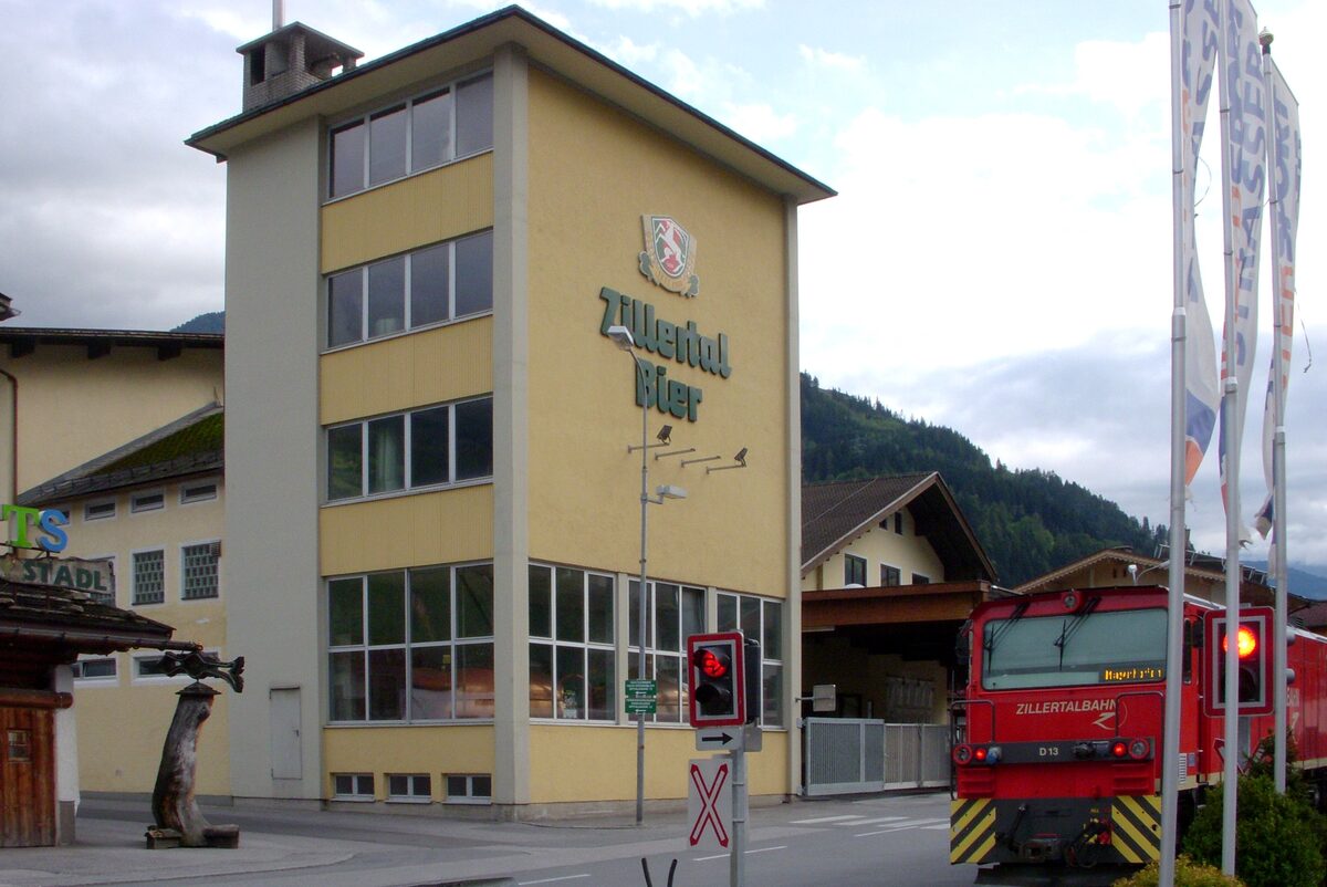 The yellow Zillertal Bier brewery building in Zell am Ziller with its beer sign on the facade, and a red Zillertalbahn train beside it