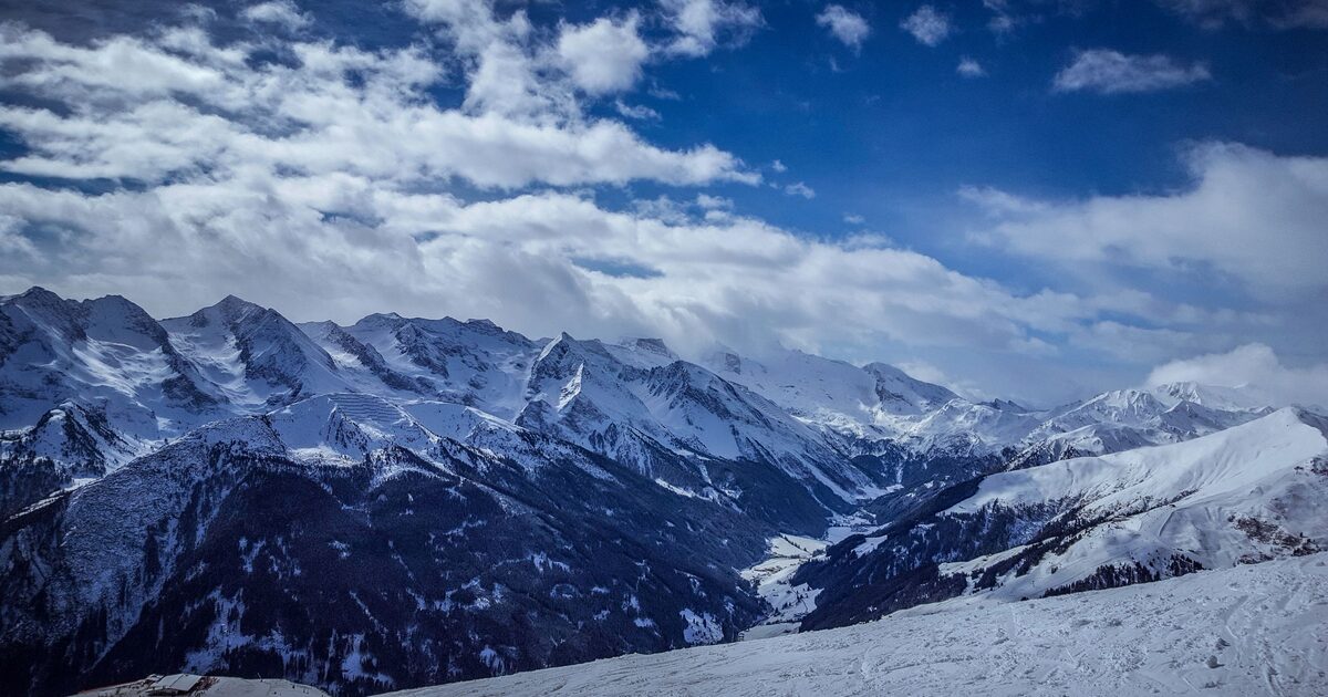 A snowy slope in the sun with the glaciated peaks of the Zillertal Alps rising behind under a blue sky and clouds