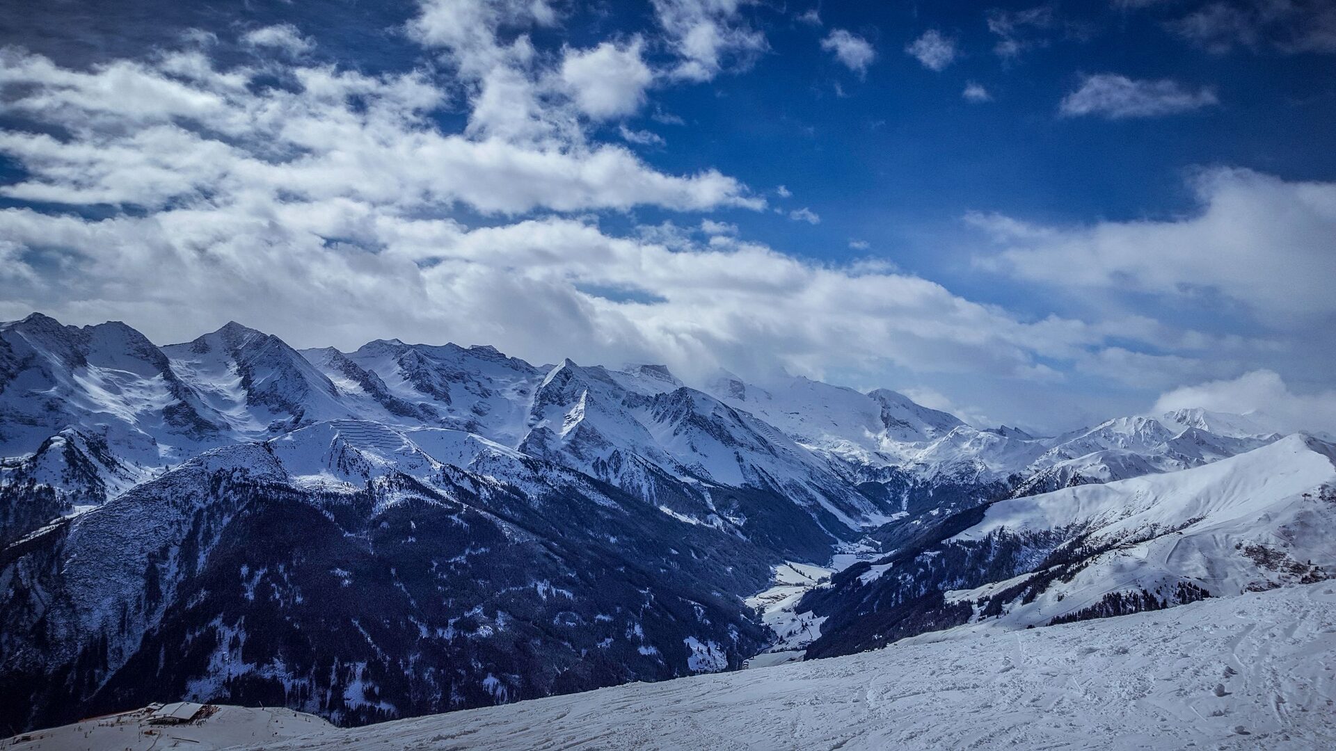 A snowy slope in the sun with the glaciated peaks of the Zillertal Alps rising behind under a blue sky and clouds