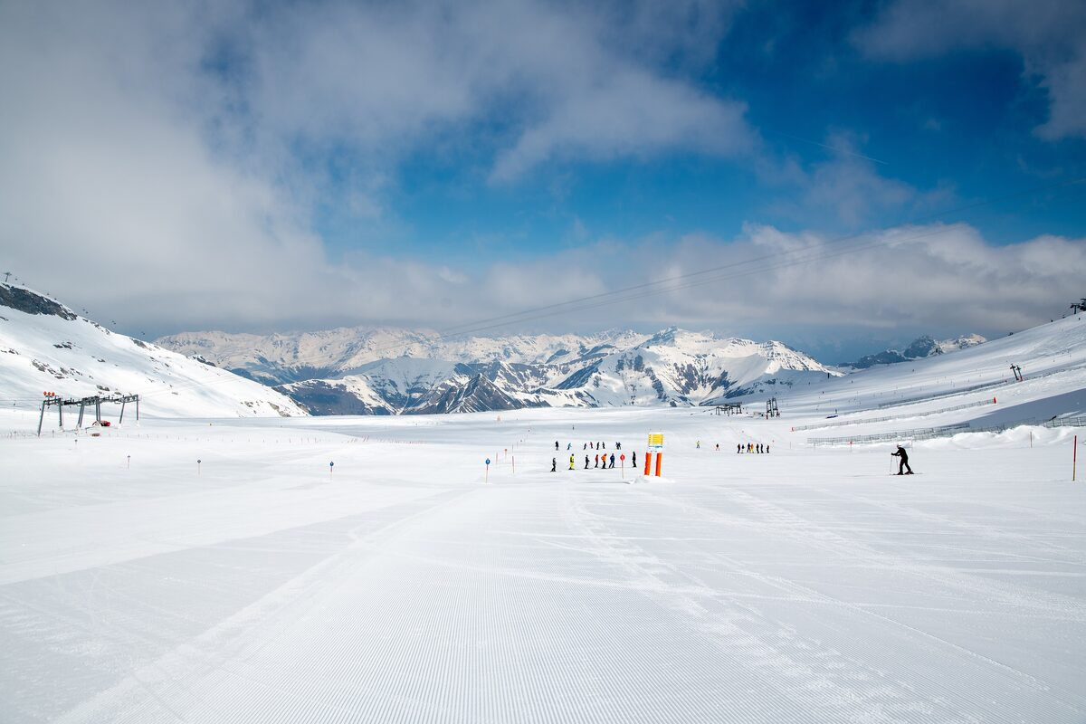 A freshly groomed piste on the Hintertux Glacier in sun, with a line of skiers in the distance and snowy peaks behind