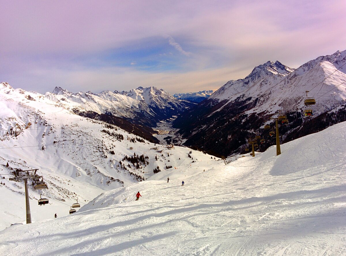 Evening light on snowy peaks above St. Anton, with a chairlift and a skier on the groomed slope below