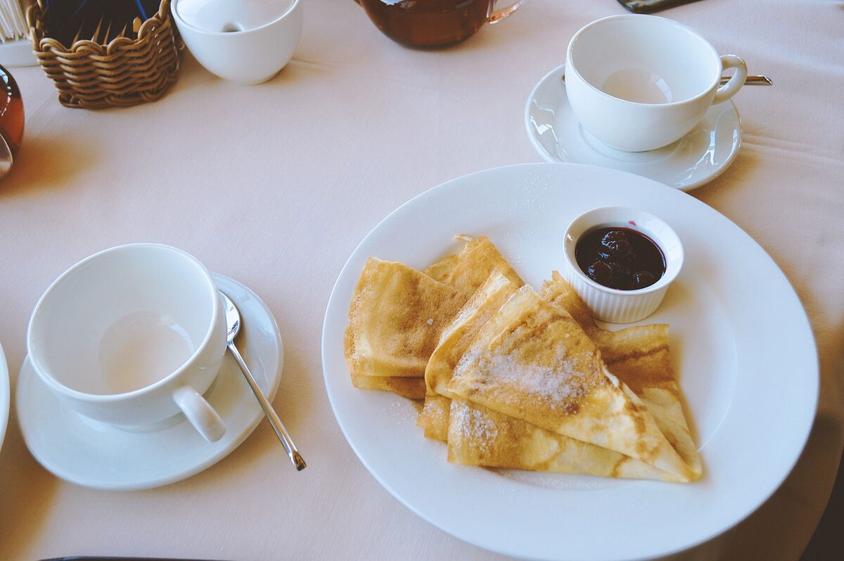 Folded crêpes dusted with sugar on a white plate, with a small pot of cherry sauce and tea cups on the table