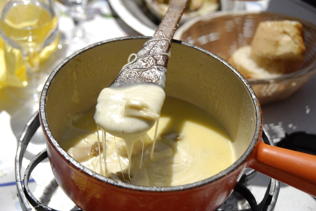 A pot of melted fondue cheese with a wooden spoon lifting a strand of cheese, and bread in a basket behind
