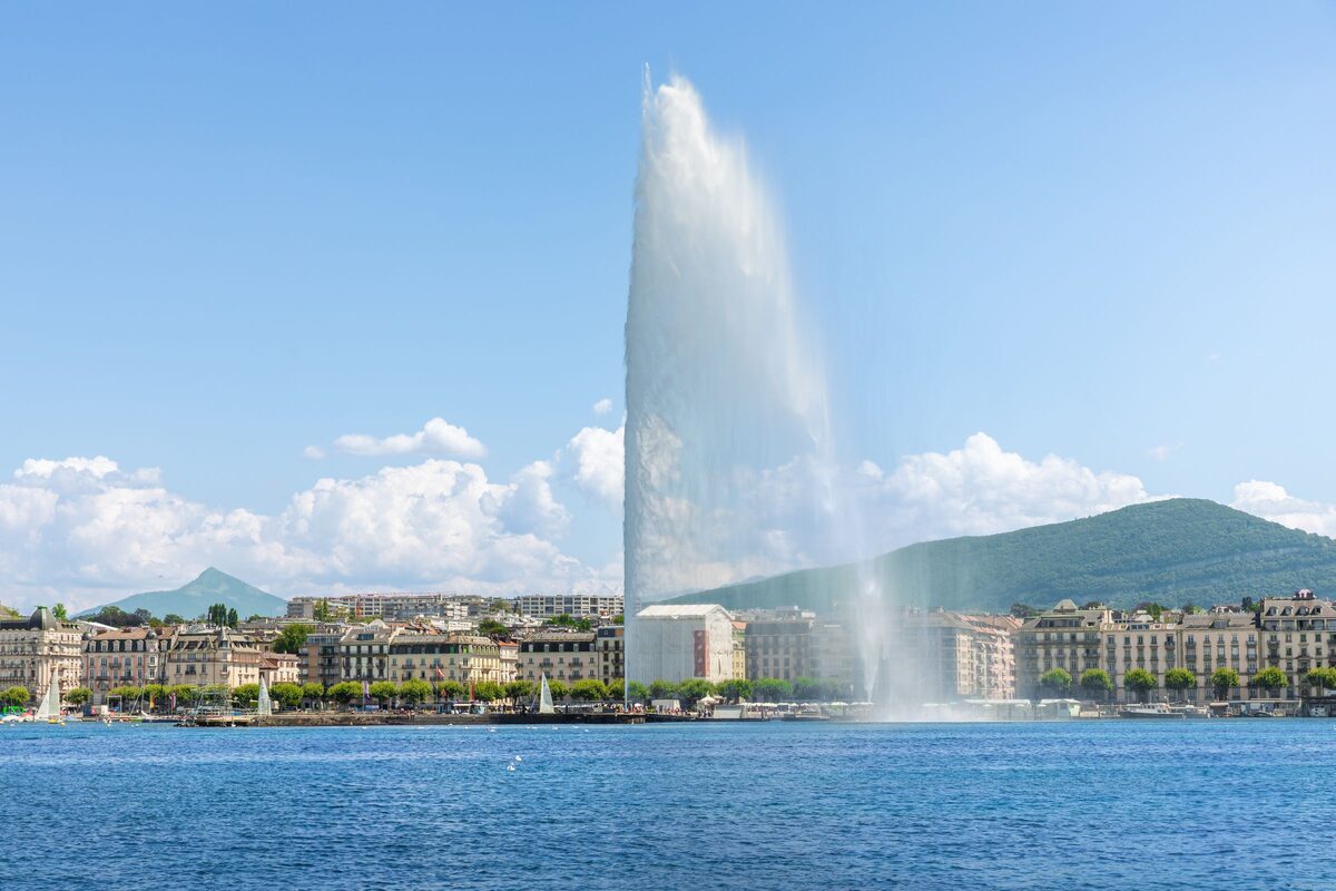 The Jet d'Eau rising from Lake Geneva on a sunny day, with the waterfront buildings and a green hill behind