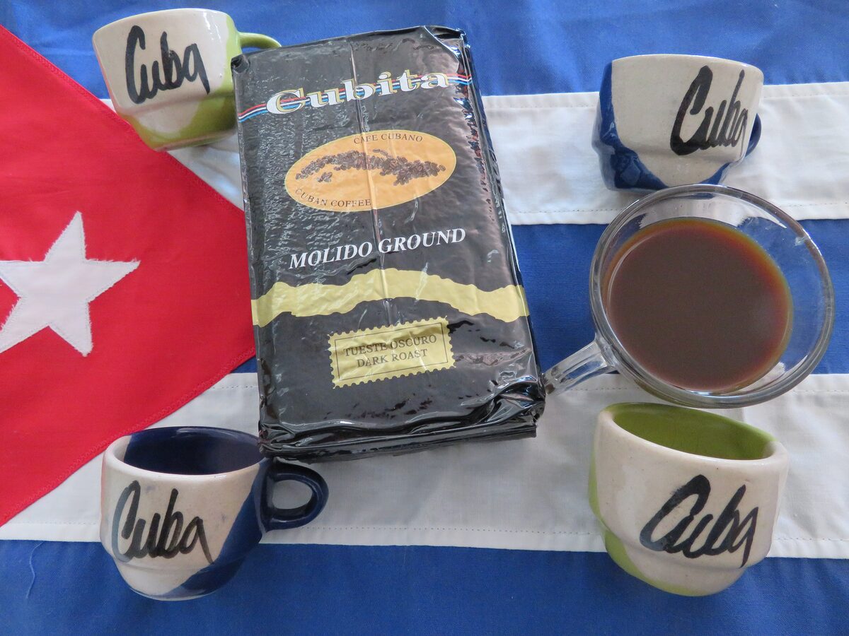 A small glass of dark Cuban cafecito among Cuba-lettered cups, a pack of ground Cuban coffee and a Cuban flag
