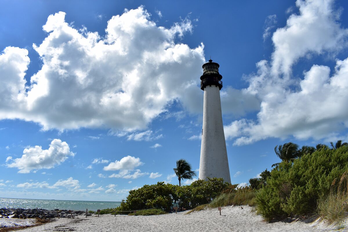The white Cape Florida Lighthouse behind a sandy beach and low green dunes, under a blue sky with big clouds
