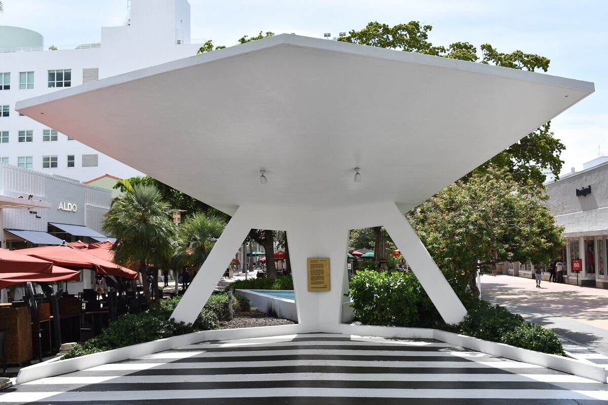 A white concrete shade canopy on Lincoln Road Mall, with palms, restaurant umbrellas and the pedestrian street beyond