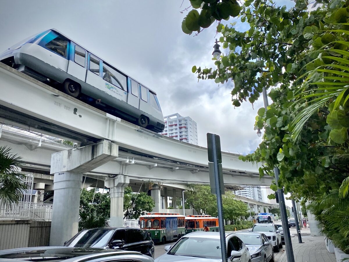 A Metromover car on its elevated concrete track in Brickell, with a trolley and an apartment tower below under a cloudy sky