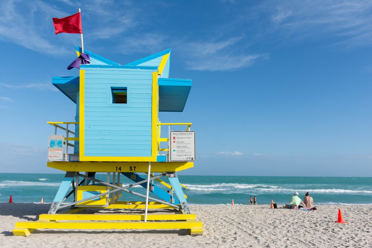 A blue and yellow lifeguard tower on the sand of South Beach, with the Atlantic and a clear blue sky behind it