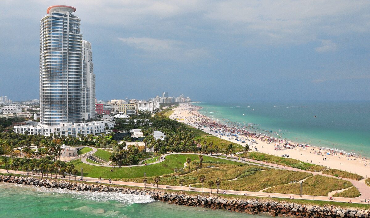 South Pointe Park from above, with its lawns and paths, the rock jetty, turquoise water and the beach of Miami Beach beside a tall tower