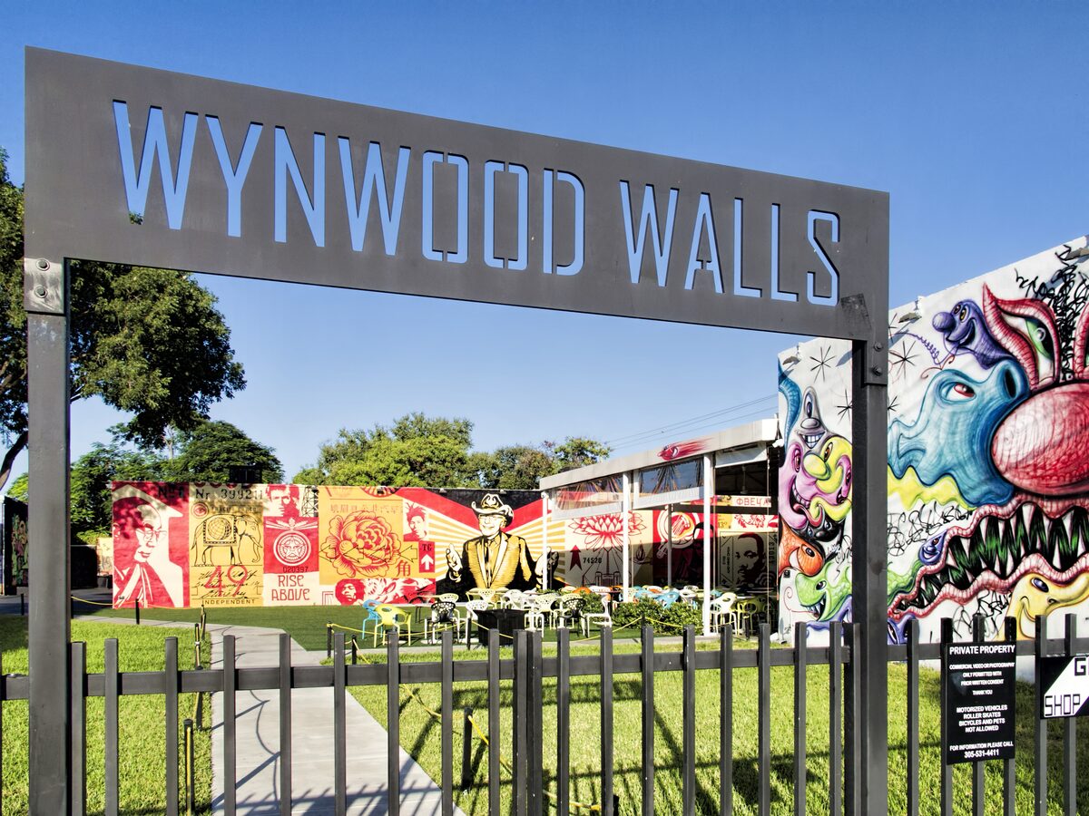 The steel Wynwood Walls entrance sign over a lawn, with colorful murals on the walls behind it