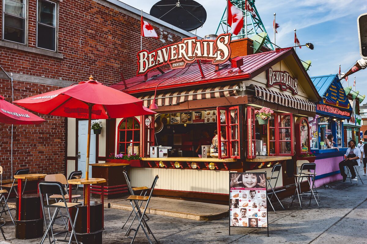 A red-roofed BeaverTails stand with its sign above the counter, umbrellas and tables in front on a brick-paved street