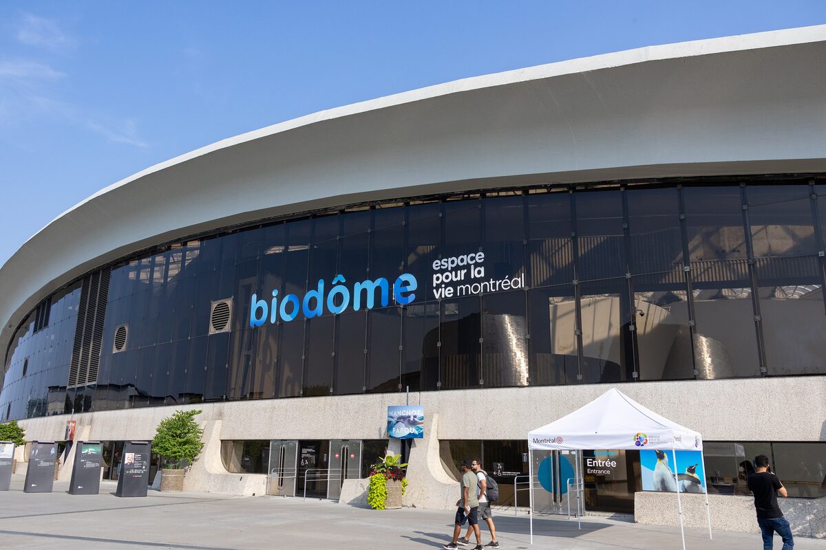 The curved front of the Biodôme building with its blue sign, visitors under a ticket tent and a clear sky above