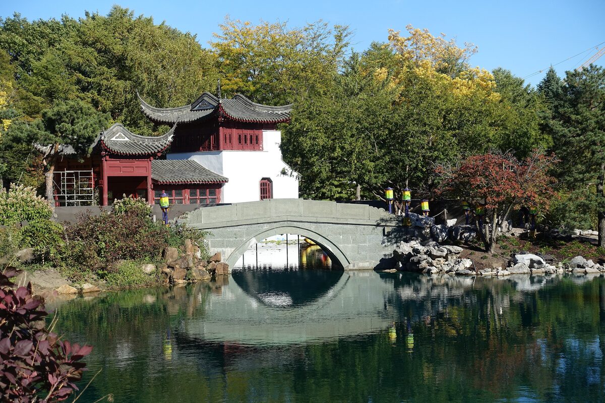 A red-roofed pavilion beside a still pond and a stone arch bridge in the Chinese Garden of the Montreal Botanical Garden