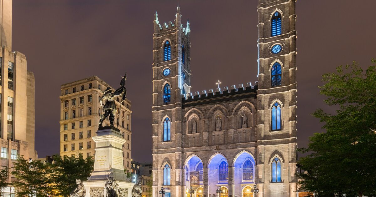 Notre-Dame Basilica lit blue at night, with its twin towers over the square and a bronze monument on the left