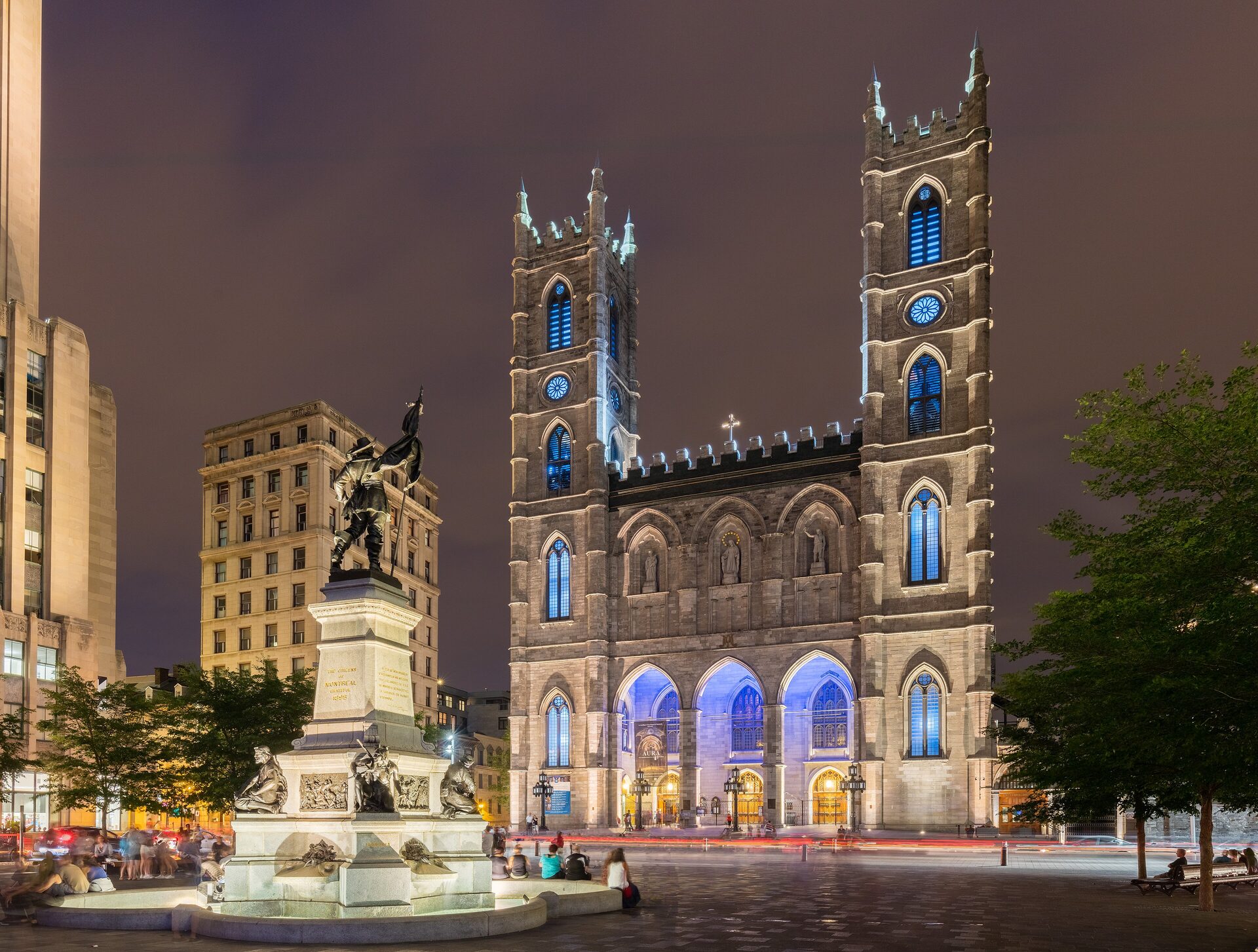 Notre-Dame Basilica lit blue at night, with its twin towers over the square and a bronze monument on the left