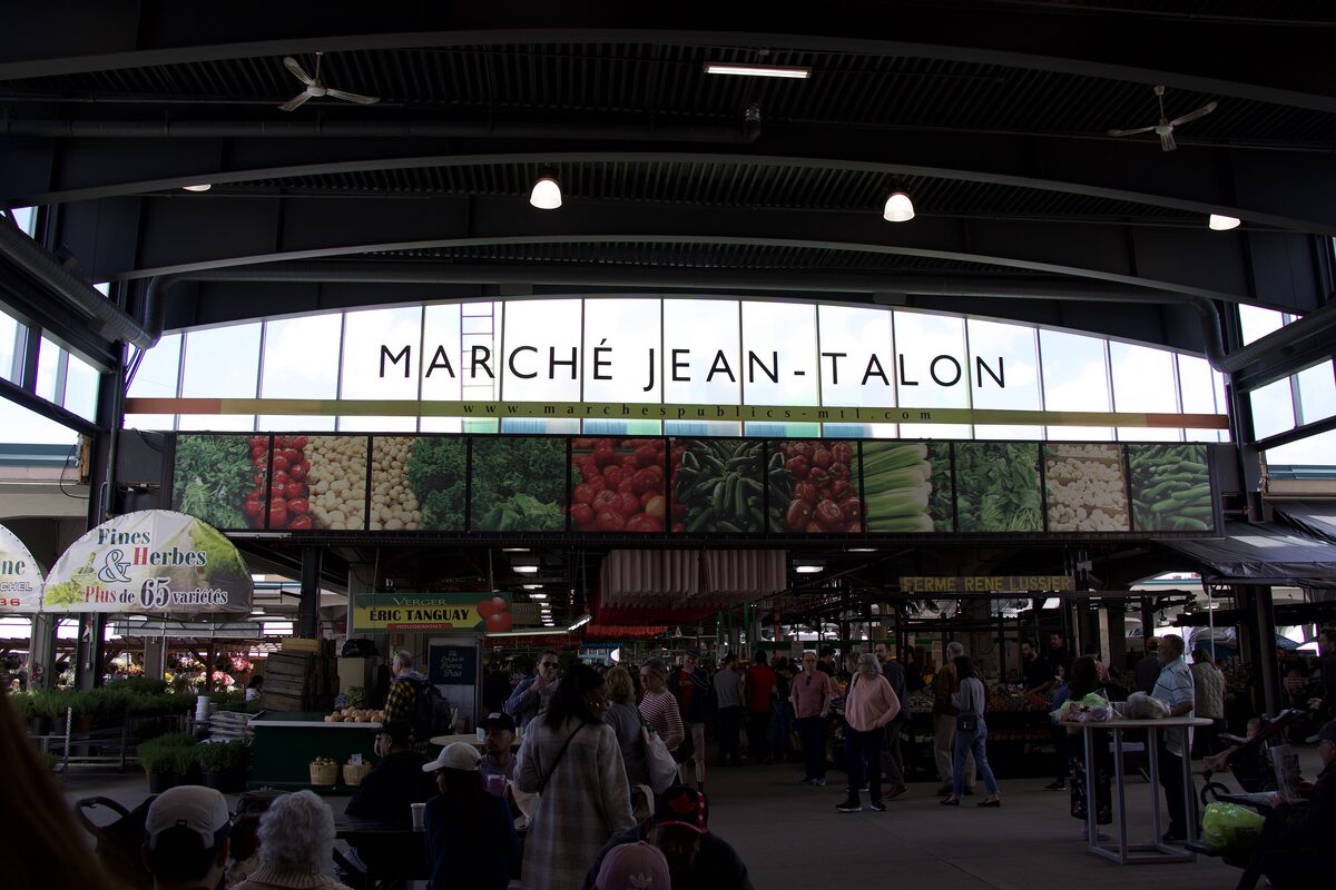 Inside Jean-Talon Market's covered hall, with the Marché Jean-Talon sign over the produce stalls and shoppers at the counters