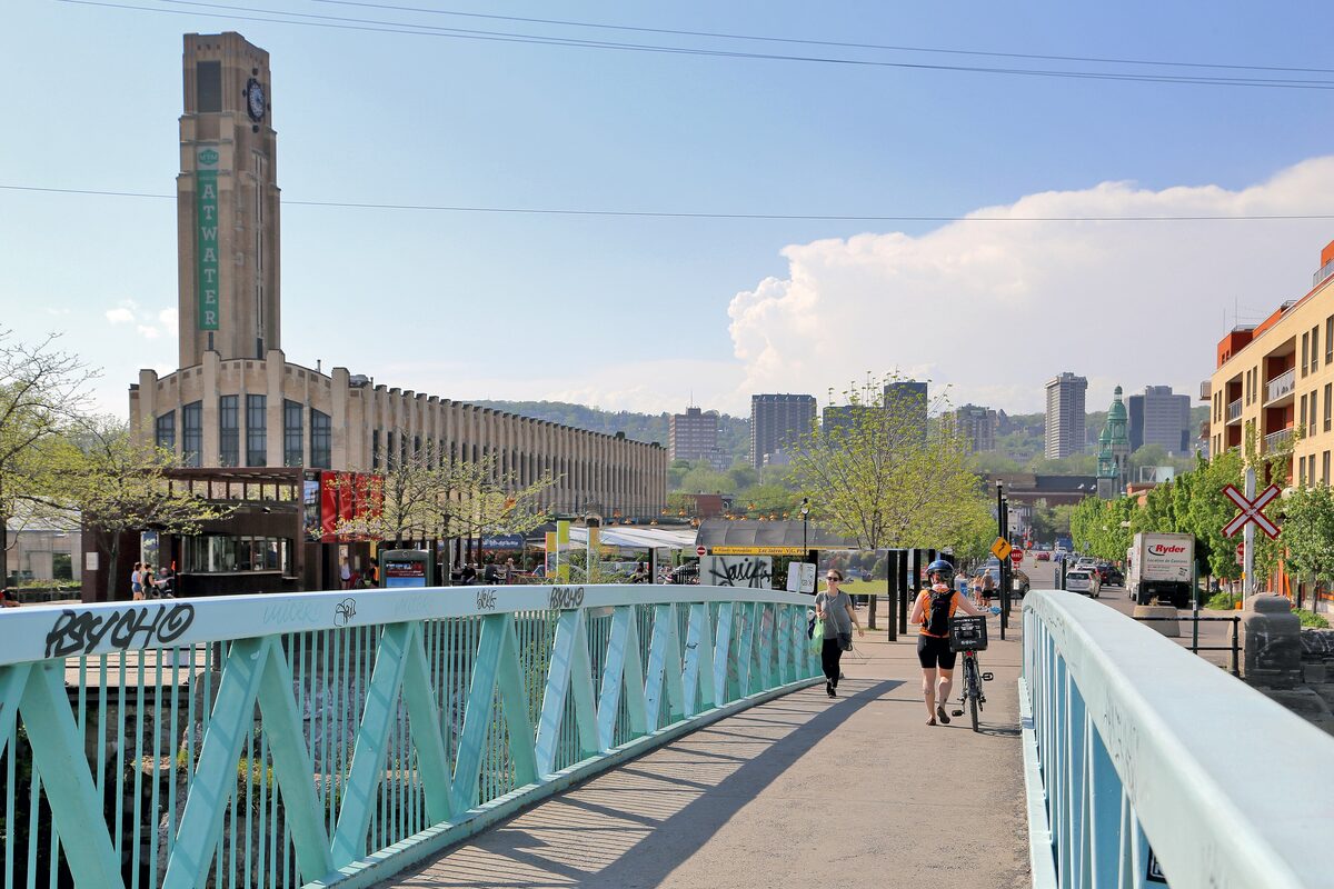 Cyclists on the Lachine Canal path crossing a turquoise footbridge, with the Atwater Market clock tower ahead