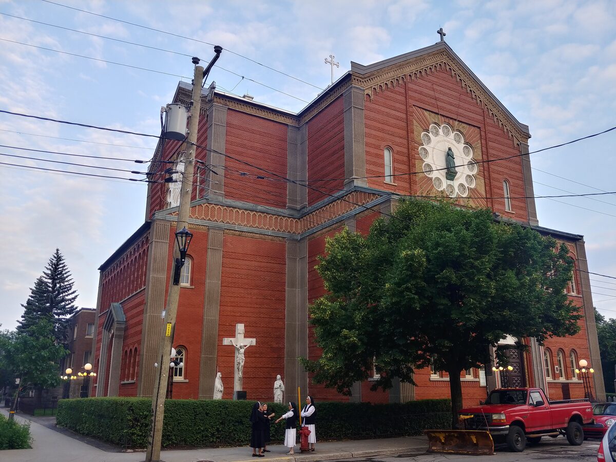 The red-brick Notre-Dame-de-la-Défense church in Little Italy, with its cross on the gable and a white cross and figures on the lawn in front