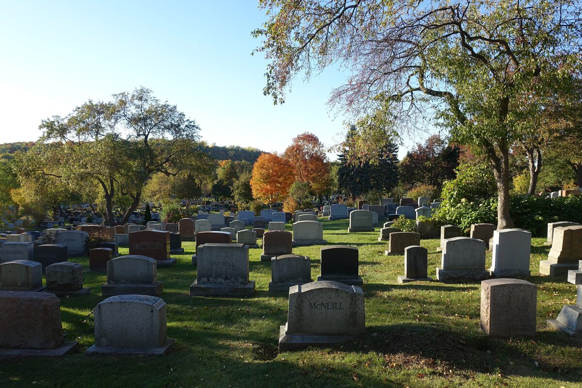 Rows of old headstones on a grassy slope in Mount Royal Cemetery, with autumn trees and a blue sky behind