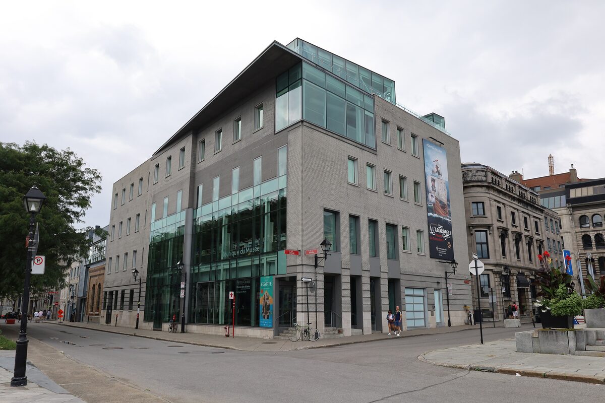 The modern glass-and-stone Pointe-à-Callière museum building on a street corner in Old Montreal, under a grey sky