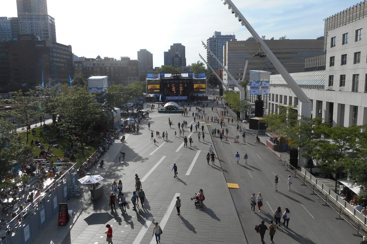 Crowds strolling across the Place des Festivals in the Quartier des spectacles during a festival, with a stage and big screen at the far end and tilted white light masts on the right