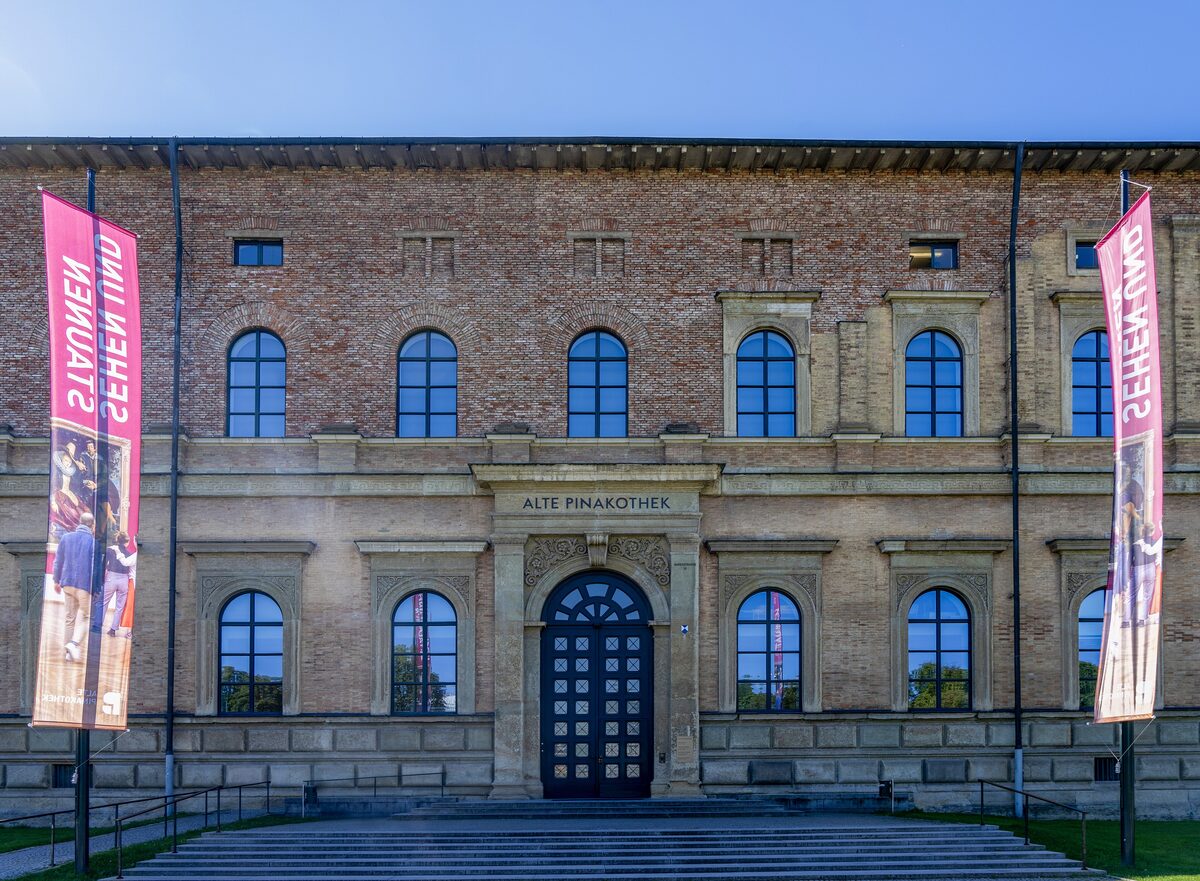 The brick facade of the Alte Pinakothek with its arched windows, main door and two banners