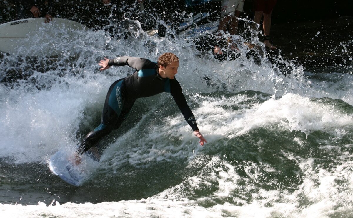 A surfer in a wetsuit leaning into the white water of the Eisbach wave
