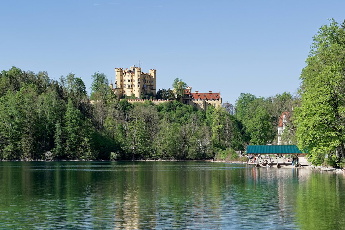 The yellow Hohenschwangau Castle on a wooded hill above the green water of the Alpsee