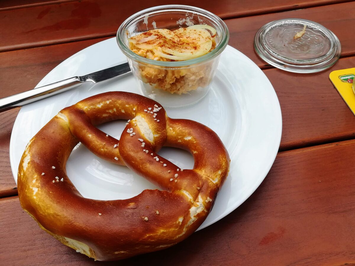 A beer garden pretzel next to a small glass jar of Obatzda topped with onion