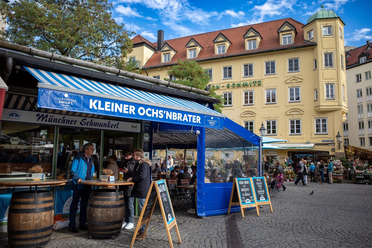 A snack stall with a blue awning and barrels on the Viktualienmarkt, with a yellow townhouse behind it
