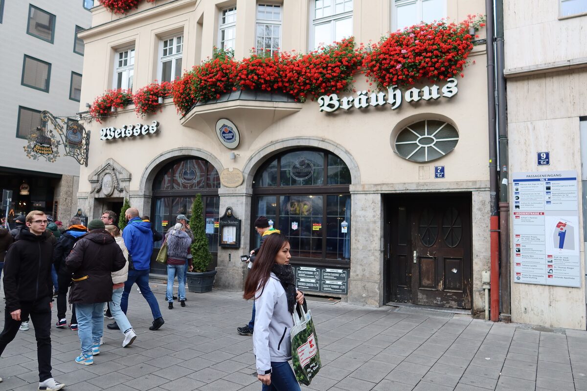 The facade of the Weisses Bräuhaus on Tal, with red geraniums on the balcony and passers-by on the pavement