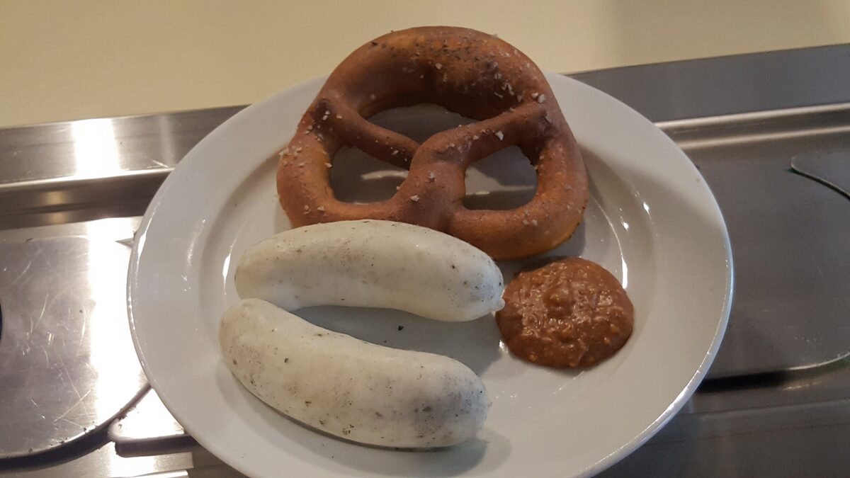 Two white veal sausages, a dark pretzel and a spoonful of sweet mustard on a plate