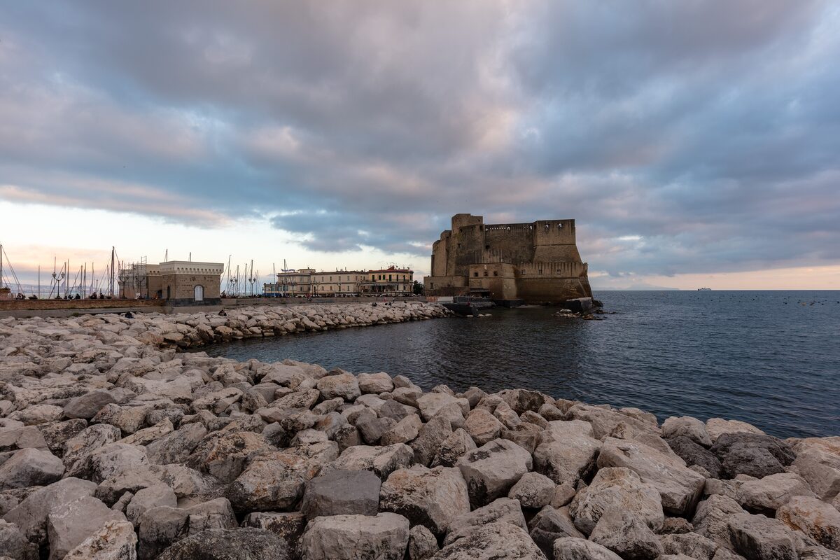 The square stone walls of Castel dell'Ovo on its islet at the end of a breakwater, under a cloudy evening sky