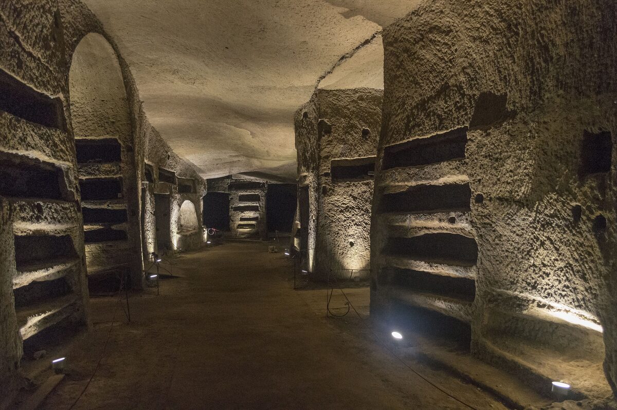 A long, softly lit gallery in the Catacombs of San Gennaro, its tuff walls lined with rows of empty burial niches