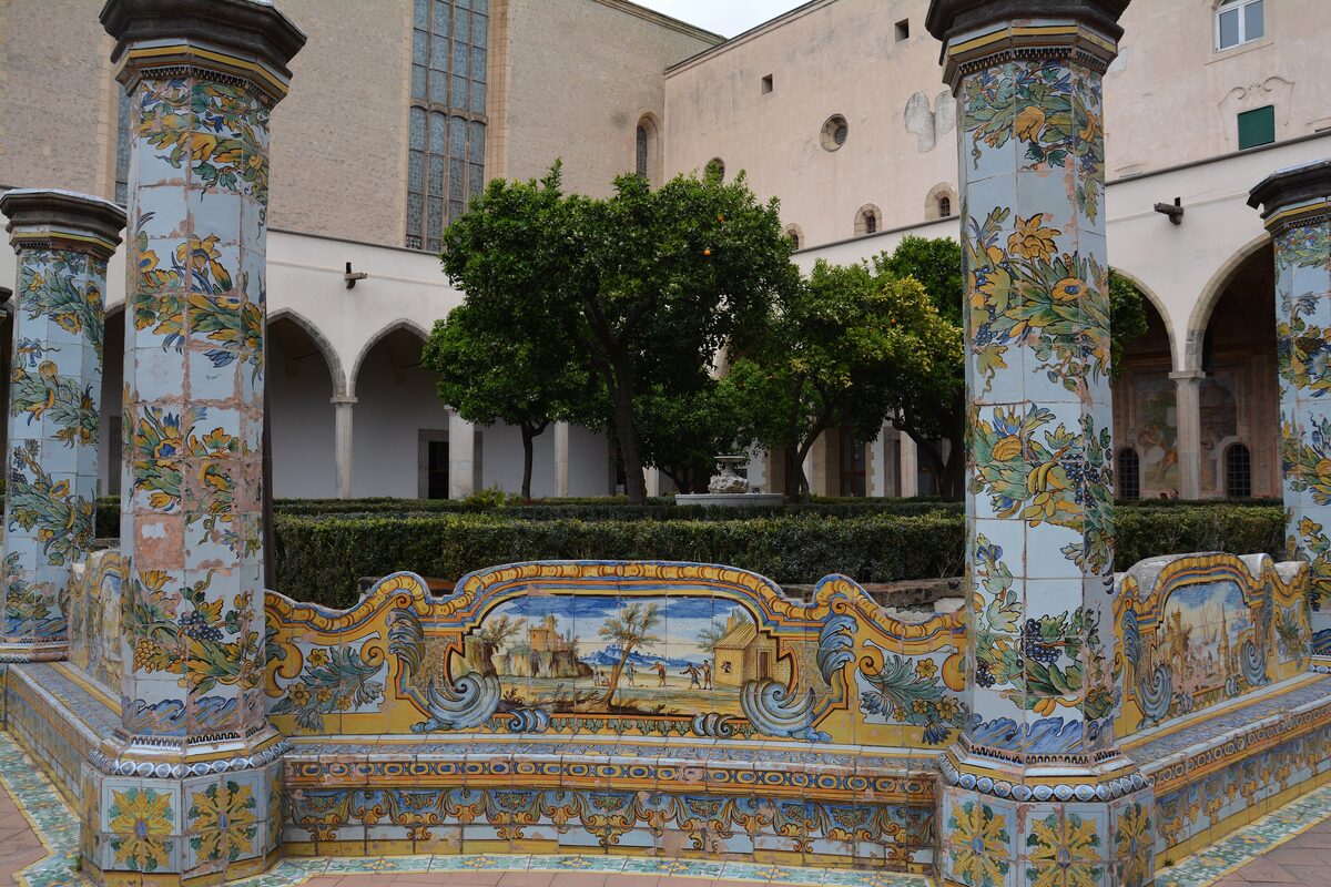 Octagonal pillars and long benches covered in yellow, blue and green majolica tiles in the cloister of Santa Chiara, with orange trees behind