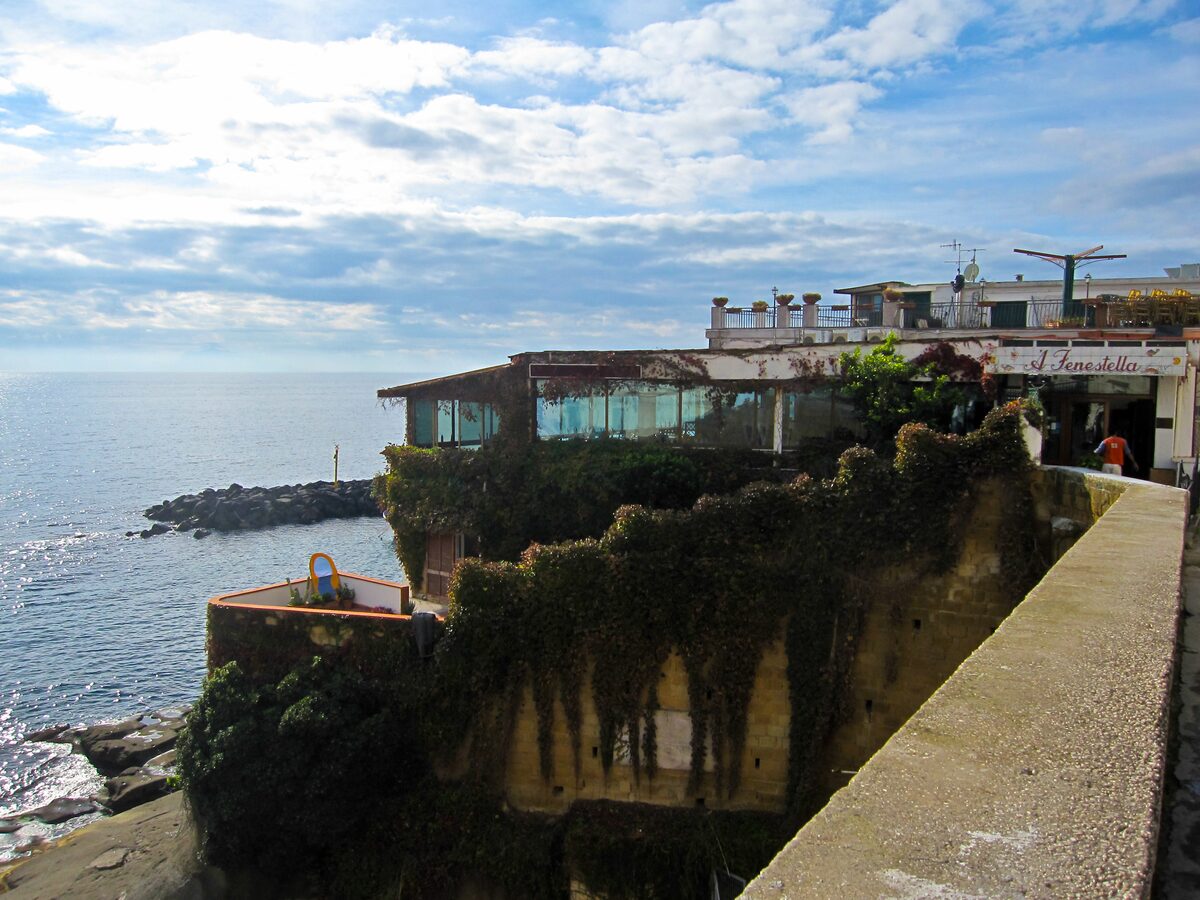 Old houses and a restaurant terrace on the rocks at Marechiaro, above a calm sea with a small breakwater