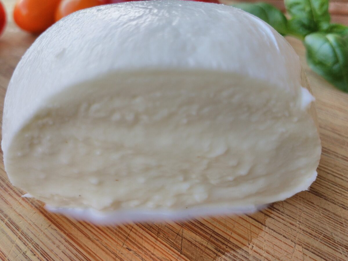 A ball of fresh buffalo mozzarella cut open on a wooden board, with tomatoes and basil behind