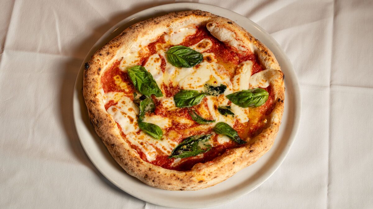 A Neapolitan margherita pizza with a puffy, charred crust, tomato, melted mozzarella and fresh basil on a white plate