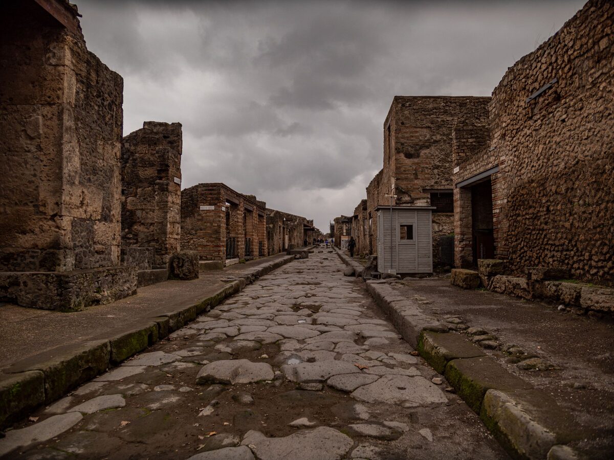 An empty paved Roman street in Pompeii, worn smooth and rutted, between the ruined walls of houses under a gray sky