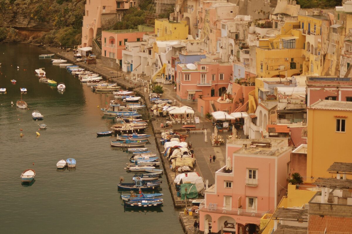 The harbor of Corricella on Procida, with pastel pink, yellow and white houses stacked above small boats moored along the quay