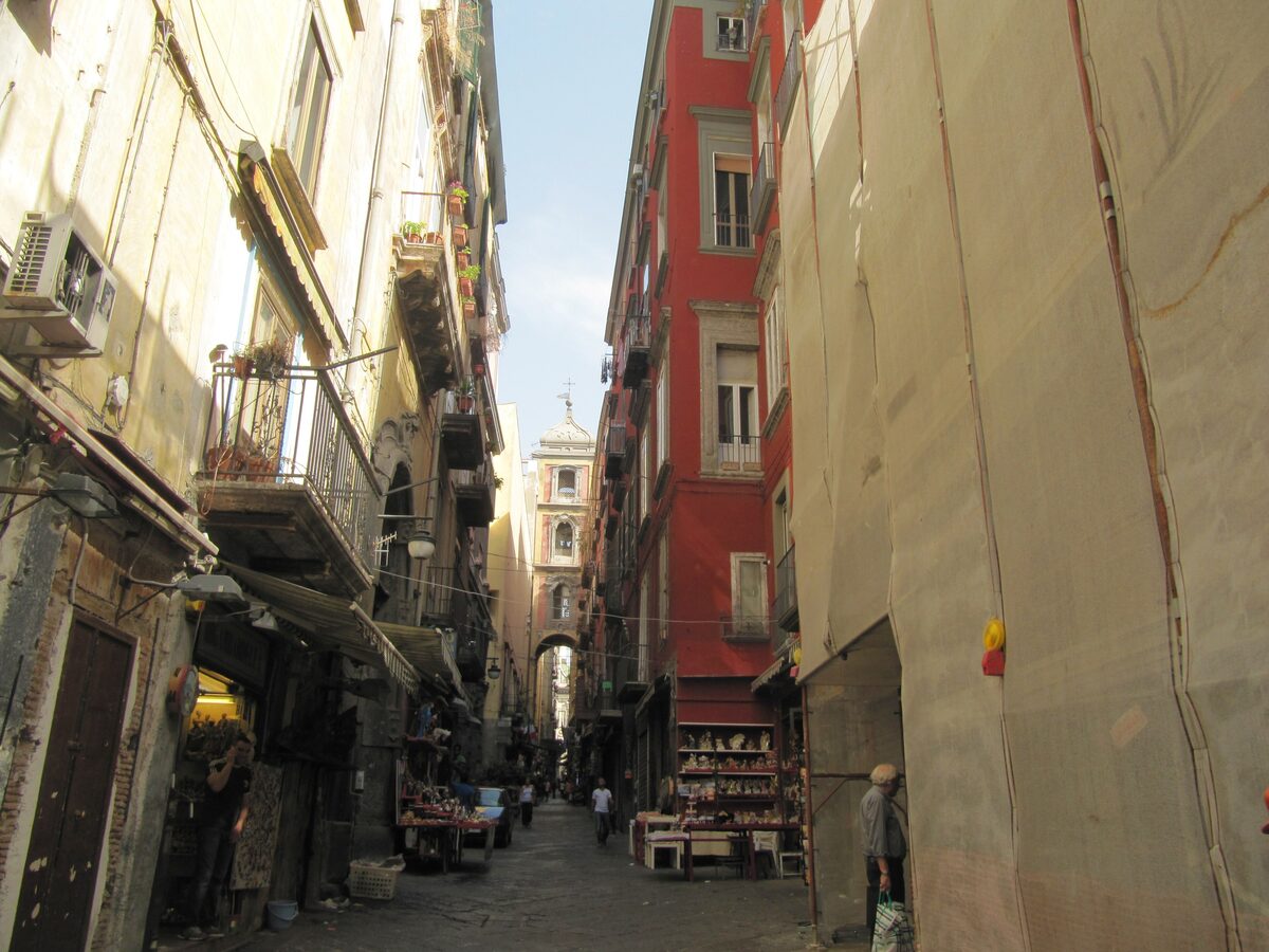 The narrow lane of Via San Gregorio Armeno with shop fronts and stalls on one side, a pink bell tower arch at the end and tall old buildings on both sides