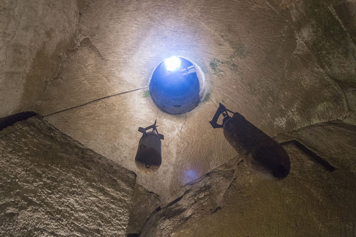A tall tuff-stone cavern under Naples lit from above through a round shaft, with two rusted World War II bombs hanging in the light