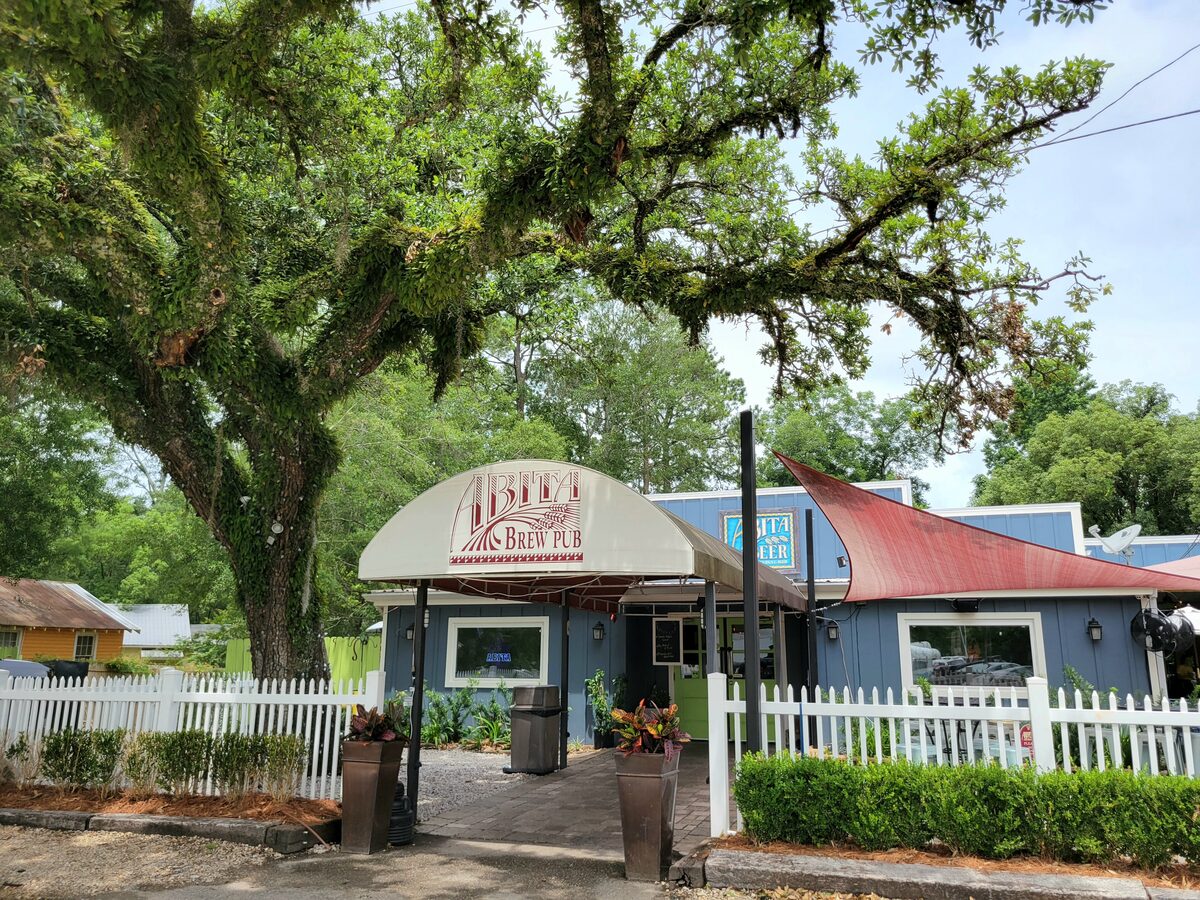 The Abita Brew Pub in Abita Springs, a blue building with a curved awning behind a white picket fence and a big live oak