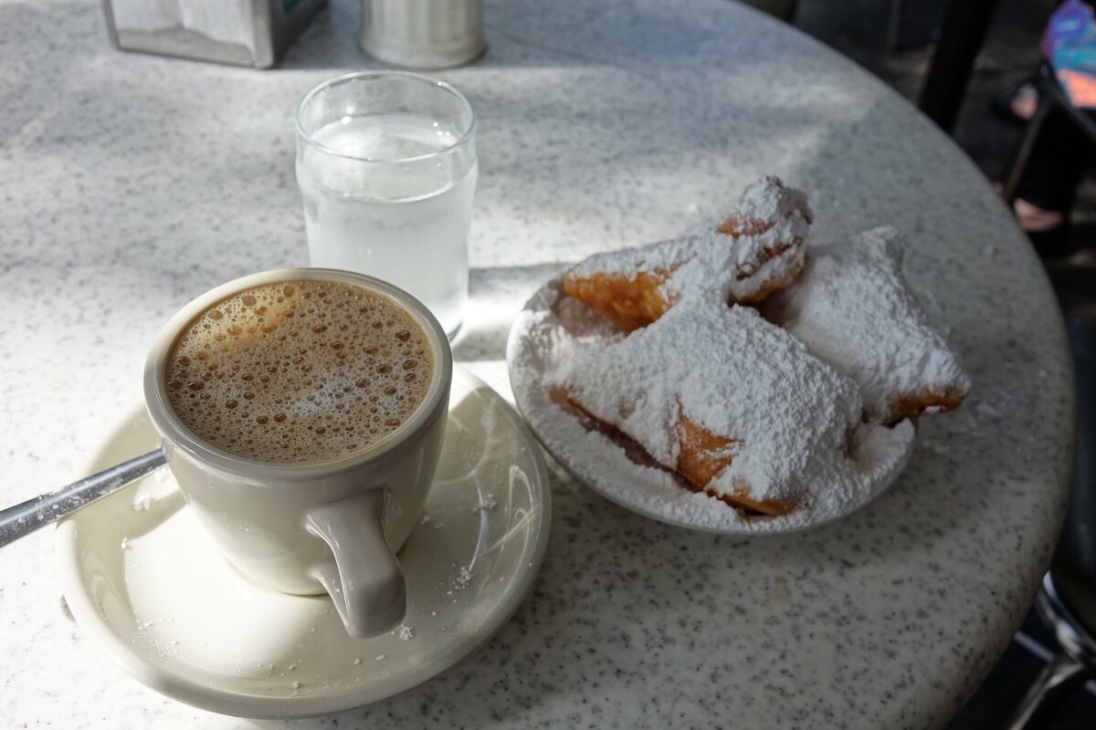 Powdered-sugar beignets on a plate beside a cup of café au lait on a cafe table