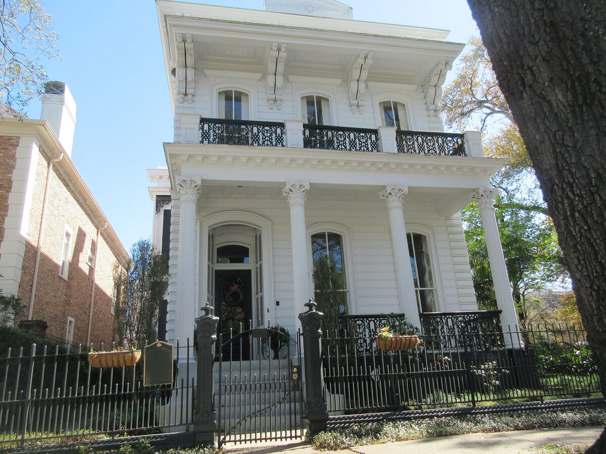 A white Greek Revival mansion with cast-iron galleries behind a fence and an old oak on St. Charles Avenue