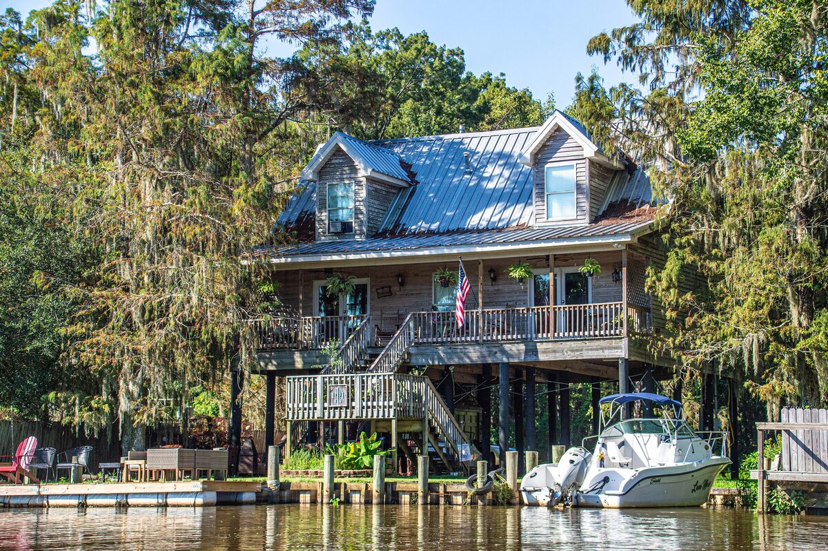 A stilted wooden house with a blue roof and a boat moored beside it on Honey Island Swamp, hung with Spanish moss