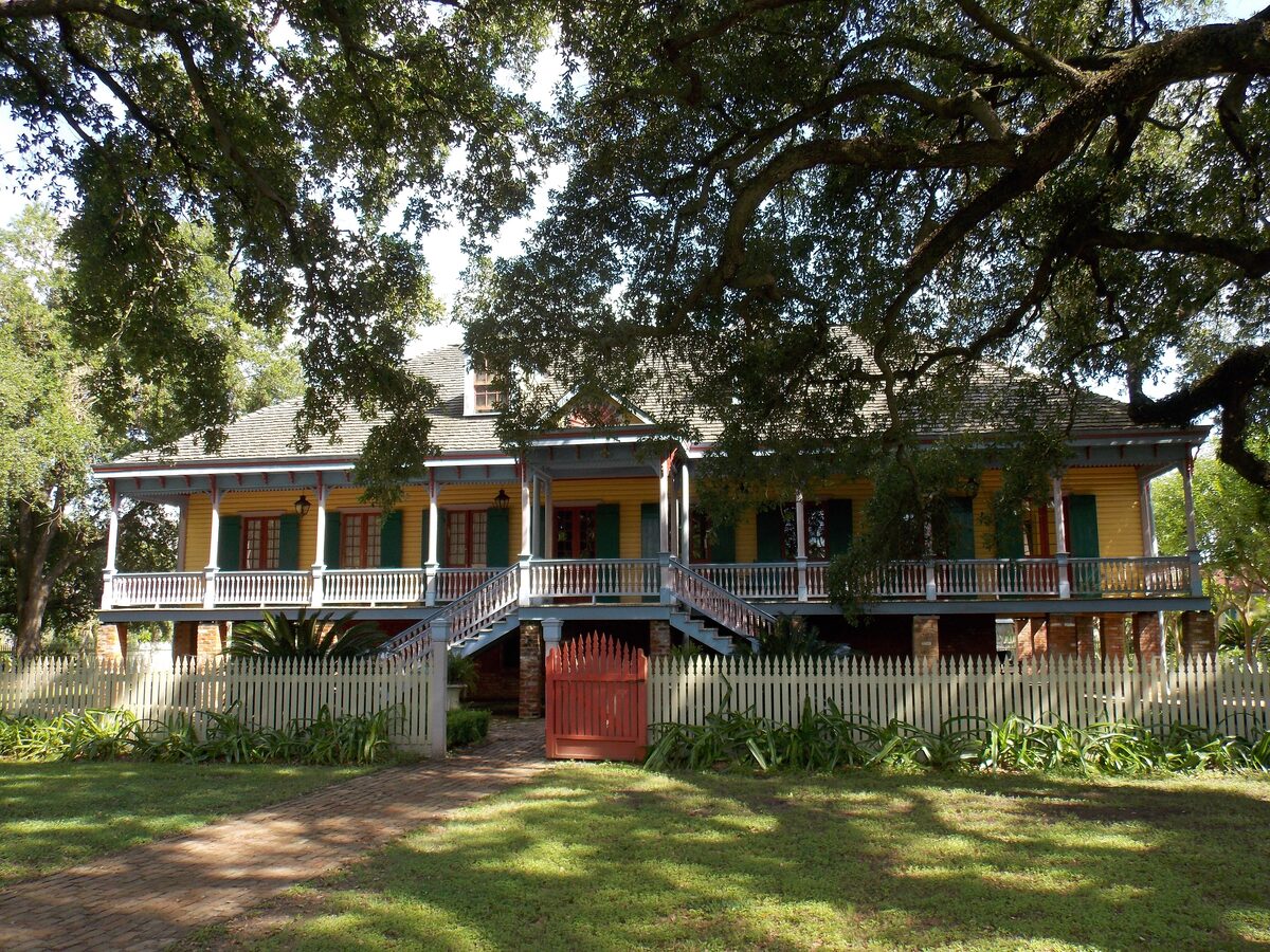 The yellow Creole big house of Laura Plantation with its raised gallery and green shutters behind a white picket fence and a red gate