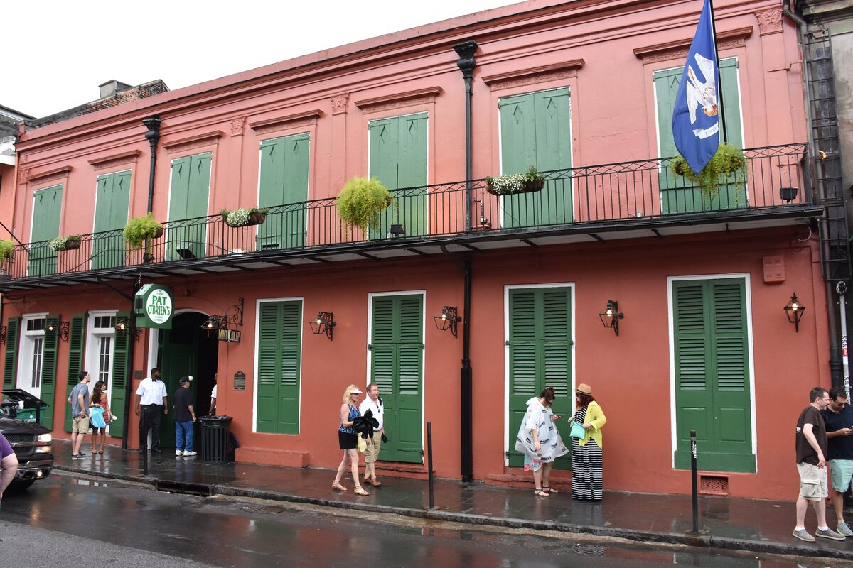 The salmon-pink two-story facade of Pat O'Brien's with green shutters and a wrought-iron balcony on a rainy street