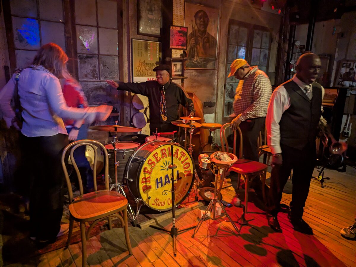 Musicians setting up on the wooden floor of Preservation Hall around a bass drum painted with the hall's name, under warm stage light